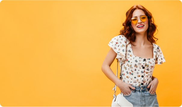 Young woman with red hair wearing a floral top and light denim jeans stands confidently against a vibrant yellow background with rounded corners. She has one hand in her pocket and carries a white purse, creating a relaxed and cheerful atmosphere.