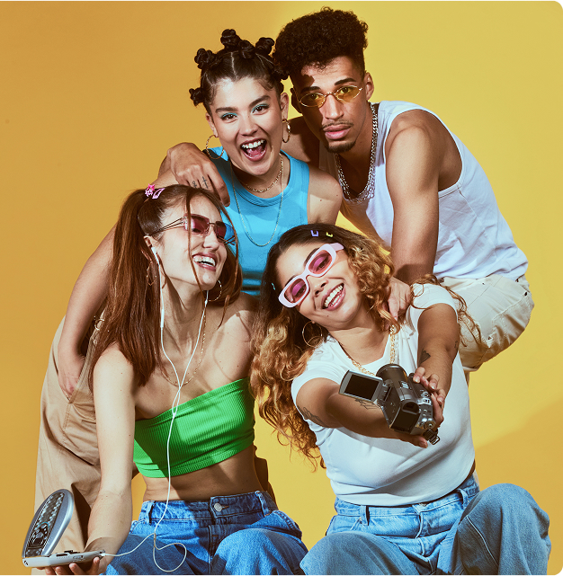 Group of four young adults wearing trendy casual clothing pose together against a solid yellow background. One person in a green top holds a portable CD player, another in a white shirt holds a video camera, and the group appears cheerful and energetic, creating a fun and lively atmosphere. No visible text in the image.