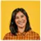 Person with shoulder-length dark hair wearing a brown and orange plaid shirt stands against a solid yellow background. The setting feels bright and cheerful, conveying a casual and friendly mood. No visible text in the image.