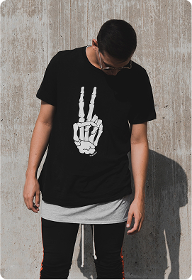 Person wearing a black t-shirt with a white skeleton hand peace sign graphic stands against a concrete wall with soft natural light. The subject has a relaxed posture, creating a casual and modern atmosphere. No visible text in the environment.