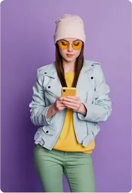 Woman standing against a purple background wearing a light blue jacket, yellow top, green pants, and a light beanie, holding a yellow smartphone with both hands. The setting feels modern and playful with a casual and relaxed mood. No visible text in the image.