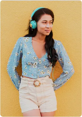Woman wearing a light blue floral long-sleeve top and high-waisted beige shorts with a decorative belt stands against a yellow textured wall. She has long dark hair and is listening to turquoise headphones. The setting feels bright and cheerful, conveying a relaxed and confident mood. No visible text in the image.