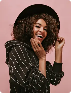 Person with curly hair wearing a black hat and a black hoodie with white pinstripes stands against a soft pink background with rounded corners. The subject is adjusting the hat with one hand and touching their hair with the other, creating a casual and stylish mood. The environment feels modern and minimalistic with a relaxed and confident tone. No visible text in the image.