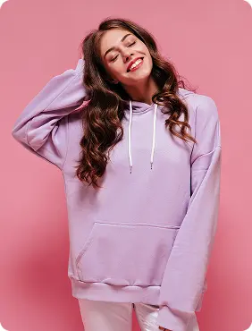 Woman with long wavy hair wearing a lavender hoodie and white pants stands against a pink background with rounded corners. She has one hand raised to her head, creating a relaxed and casual mood. The environment feels modern and playful with soft lighting and no visible text in the image.