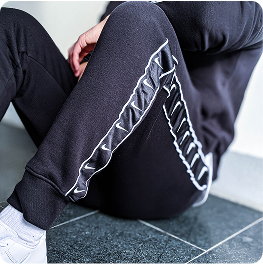 Person wearing black joggers with white piping detail sits on a tiled floor with one knee raised and hand resting on the leg. The environment is modern and minimalistic with a calm and relaxed mood. No visible text in the image.