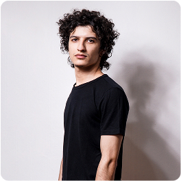 Man with curly hair wearing a black t-shirt stands in a minimalistic studio with a white wall and soft lighting. His posture is relaxed and casual, creating a calm and modern atmosphere. No visible text in the image.