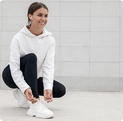 Person wearing a white hoodie and black leggings crouches down to tie their white sneakers on a light tiled outdoor surface with a modern gray stone wall in the background. The scene feels calm and minimalistic with a focus on casual athletic style. No visible text in the image.