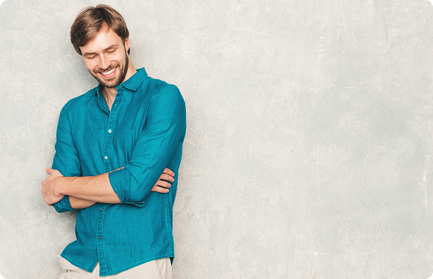 Man wearing a teal button-up shirt with sleeves rolled up stands against a textured light gray wall with rounded corners. His arms are crossed and his posture is relaxed, creating a calm and confident mood. The setting is minimalistic and modern with no visible text in the image.