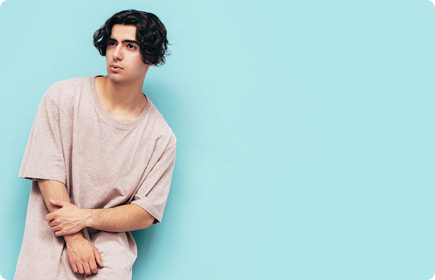 Young man with wavy dark hair wearing an oversized light beige t-shirt stands against a pastel blue background with rounded corners. He is leaning slightly to one side with his arms crossed in a relaxed and casual pose. The setting feels modern and minimalistic, conveying a calm and approachable mood. No visible text in the image.